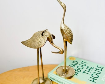 Op de afbeelding: Twee messing kraanvogelbeeldjes, één met de kop naar beneden en de ander rechtop. De vogels hebben gedetailleerde veermotieven en zijn gemonteerd op ronde voetstukken. Een boek met de titel "Mad About The House" is zichtbaar.