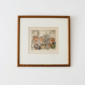May include: A framed print featuring a still life illustration of various kitchen items including terracotta jars, glass containers, a bowl with fruit, and dried plants. The artwork is enclosed in a square wooden frame with a cream-coloured mat board.