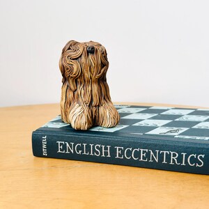May include: A brown ceramic dog figurine with detailed fur. The dog sits on a book titled "ENGLISH ECCENTRICS". The book has a blue cover with a black and white checkered pattern.