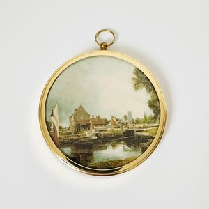 May include: A round, framed watercolor painting depicting a waterside scene with buildings, a bridge, and a church steeple. The painting is set within a gold-colored metal frame with a small ring at the top for hanging.