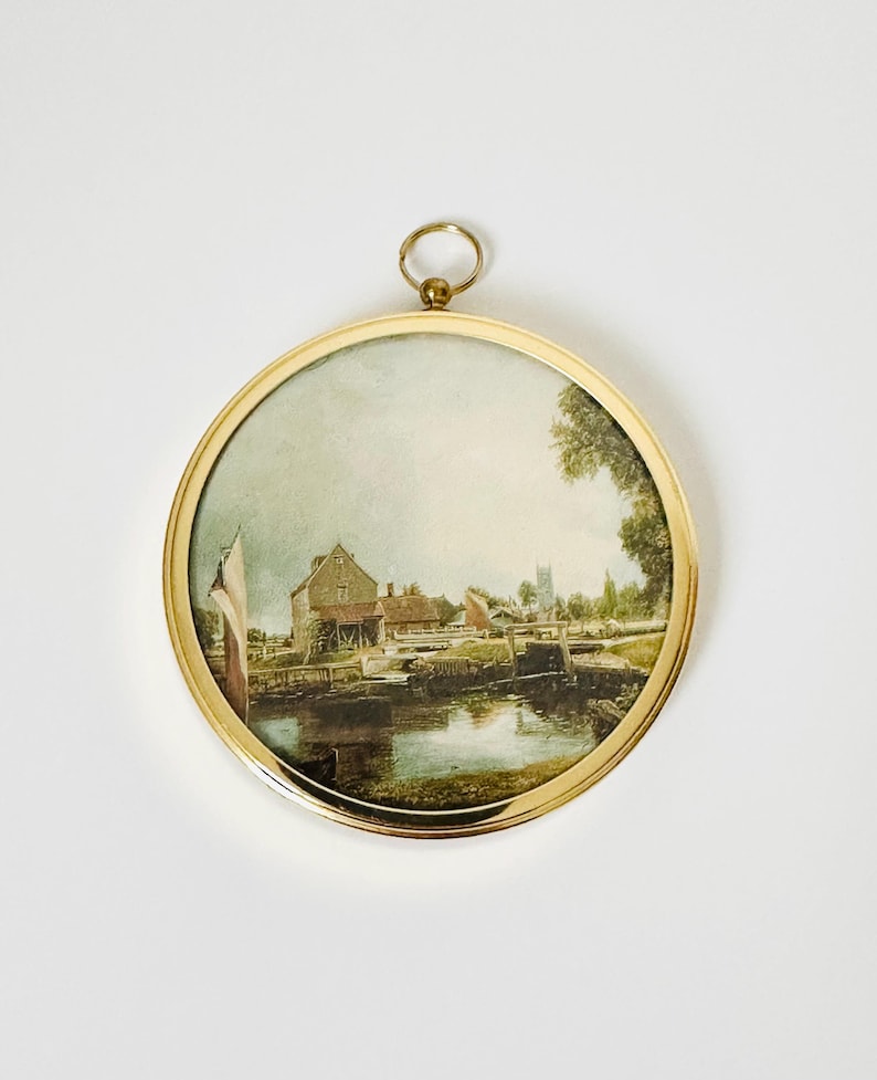 May include: A round, framed watercolor painting depicting a waterside scene with buildings, a bridge, and a church steeple. The painting is set within a gold-colored metal frame with a small ring at the top for hanging.