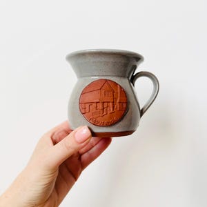May include: A gray ceramic mug with a handle and a round, reddish-brown emblem featuring a house design. The word "Mousehole" is inscribed below the house. The mug has a wide mouth and a curved handle.
