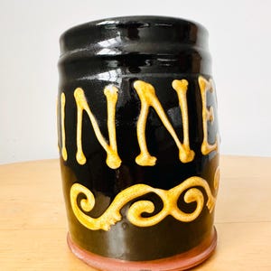 May include: A black ceramic jar with the word "WINNER" in gold lettering. The jar has a gold decorative swirl design near the bottom and a reddish-brown base. The jar is cylindrical and has a glossy finish.