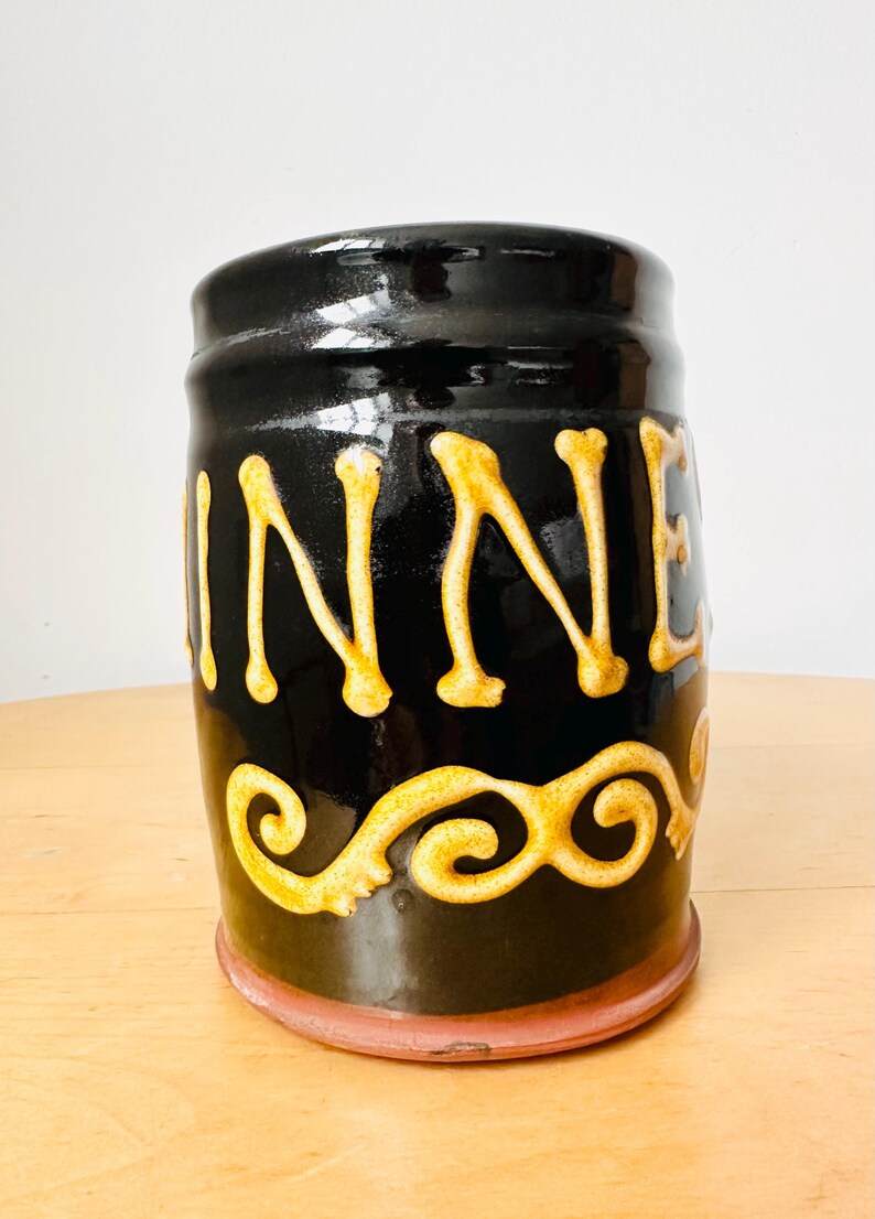 May include: A black ceramic jar with the word "WINNER" in gold lettering. The jar has a gold decorative swirl design near the bottom and a reddish-brown base. The jar is cylindrical and has a glossy finish.