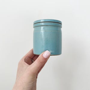 以下が含まれることがあります： 蓋付きの小さな円筒形の水色の陶器の瓶。瓶は斑点のある質感で、蓋には濃い茶色の縁があります。白い背景を背景に、人の手で瓶が持たれています。