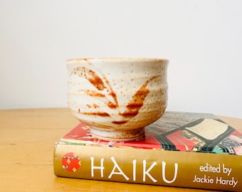 Puede incluir: Un pequeño cuenco de cerámica blanquecina con diseños marrones en forma de hojas. El cuenco está sobre un libro titulado "HAIKU" editado por Jackie Hardy. El libro tiene una cubierta roja con letras doradas y elementos decorativos.
