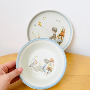 May include: Two white ceramic plates with a blue rim. The plates feature a whimsical illustration of a child with a kite and a child with a dog. The plates have a vintage aesthetic.