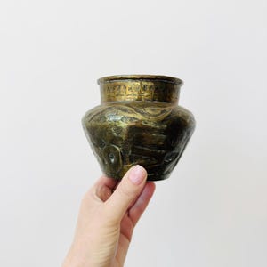 May include: A small, dark brass vessel with a wide, rounded body and a short, flared neck. The surface features intricate, etched patterns and a weathered, aged patina, showcasing a textured finish across the exterior of the metal container.