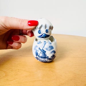 May include: A ceramic duckling figurine in white and blue, emerging from a cracked eggshell. The eggshell features a blue windmill scene. The duckling has blue eyes and a blue beak. The figurine is on a light brown surface.