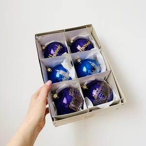May include: A set of six Christmas baubles in a box. The baubles are blue and purple with gold detailing. The box is beige and the baubles are presented in white paper.