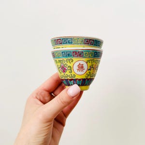 May include: Two small, yellow, decorative ceramic cups with intricate floral and geometric patterns in red, green, and blue. Each cup has a Chinese character in a red circle. The cups are stacked.