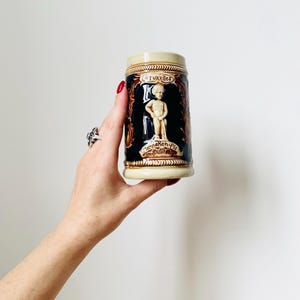 Op de afbeelding: Een keramische bierkan met een bruin en wit ontwerp. De kan heeft een afbeelding van een kleine jongen die in een fontein plast. De tekst "Bruxelles" en "Manneken-Pis" is zichtbaar op de kan.