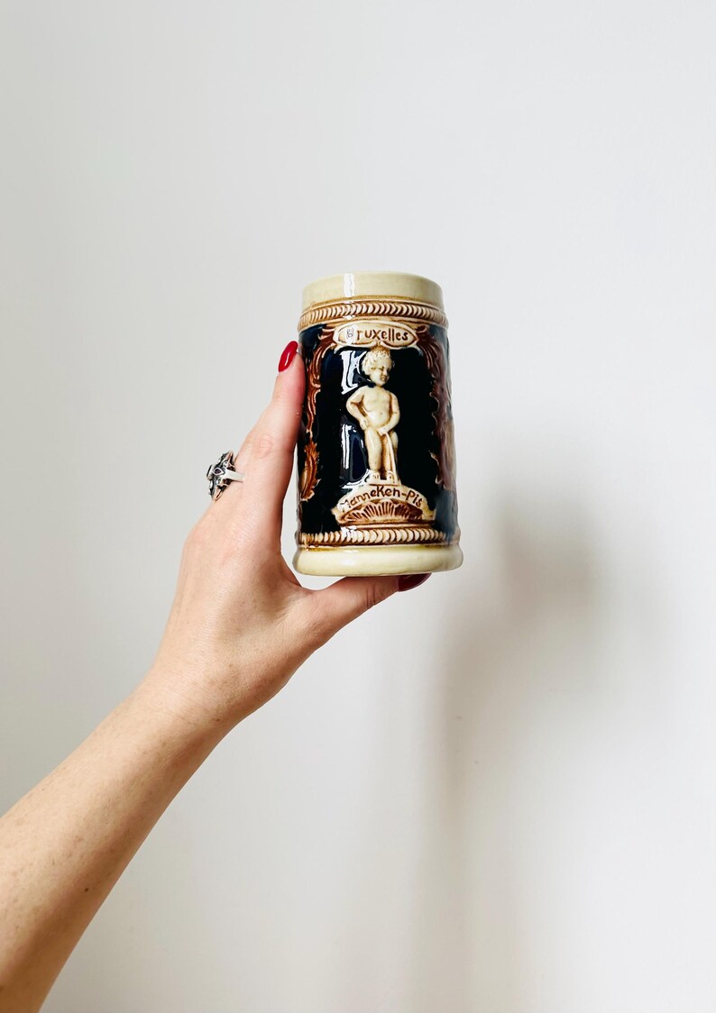 Op de afbeelding: Een keramische bierkan met een bruin en wit ontwerp. De kan heeft een afbeelding van een kleine jongen die in een fontein plast. De tekst "Bruxelles" en "Manneken-Pis" is zichtbaar op de kan.