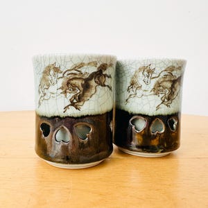 Puede incluir: Dos tazas cilíndricas de cerámica con acabado de esmalte craquelado. La parte superior es de color verde claro con ilustraciones de caballos en marrón oscuro, mientras que la inferior es marrón oscuro con tres recortes en forma de corazón.