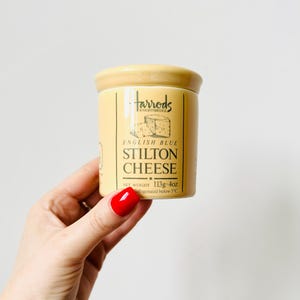 May include: A light yellow ceramic jar of Harrods English Blue Stilton Cheese. The jar has a lid and a label with the Harrods logo and cheese information. The net weight is 4oz. The jar is held by a hand with red nail polish.