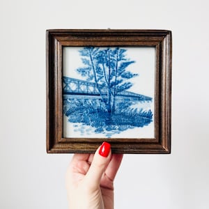 May include: A framed blue and white tile featuring a bridge and trees. The artwork is set in a dark brown wooden frame. The tile has a classic, vintage aesthetic, with a detailed landscape scene.