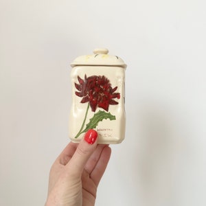 Könnte beinhalten: Eine kleine weiße Keramikdose mit Deckel, verziert mit einer handgemalten roten Blume mit grünen Blättern. Die Dose trägt den Schriftzug "WARRATAH N.S.W." auf der Vorderseite.