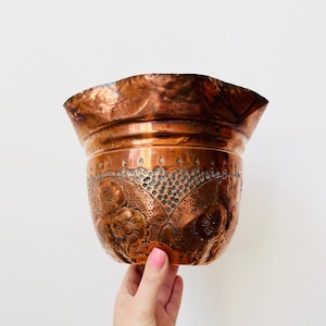 May include: A copper planter with a flared rim and floral and dot pattern. The planter is a warm, reddish-brown colour. The floral design is etched into the metal. The planter is held by a person.