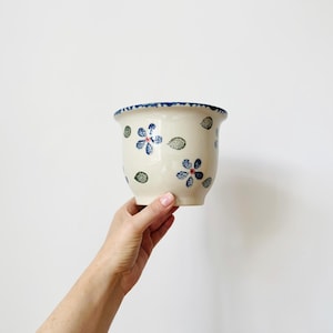 May include: A cream-coloured ceramic planter featuring a hand-painted pattern of small blue flowers with red centres and green leaves. The rim of the pot is decorated with a blue speckled border, and the overall shape is rounded with a flared top edge.