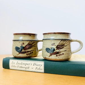 Puede incluir: Dos tazas de cerámica beige con bordes y asas marrones. Cada taza presenta un diseño pintado a mano con formas abstractas marrones y azules. Las tazas están colocadas sobre un libro verde titulado "An Innkeeper's Diary".