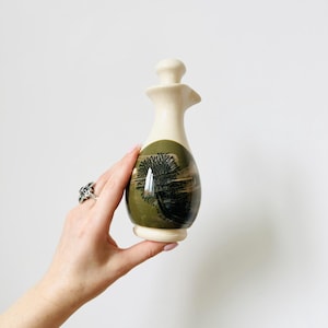 Könnte beinhalten: Eine Keramikölflasche mit cremefarbenem Oberteil und Boden sowie einem grün-braunen bemalten Design auf dem Körper. Die Flasche hat einen Ausguss und einen kleinen Verschluss. Das Design scheint eine Landschaft oder ein Baum zu sein.