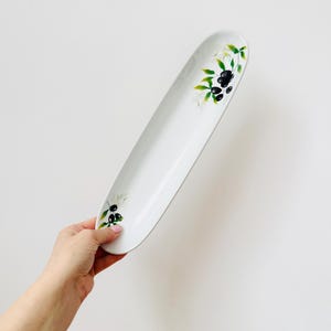 May include: A white, oval serving platter with a painted olive branch design. The platter is held by a hand and features black olives and green leaves. The design is repeated at both ends of the platter.