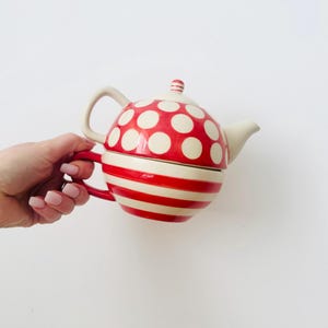 Op de afbeelding: Een rood-witte theepot met stippen met een bijpassend deksel en een rood-witte gestreepte basis. De theepot heeft een crèmekleurige handgreep en tuit. Het ontwerp is grillig en decoratief.