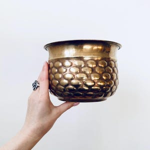 Op de afbeelding: Een vintage messing plantenbak met een getextureerd, kuiltjesoppervlak. De plantenbak heeft een brede, afgeronde vorm en een gladde rand. Het messing heeft een warme, gouden tint. De plantenbak wordt vastgehouden.