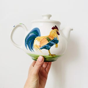 May include: A white ceramic teapot featuring a hand-painted rooster with yellow, blue, and red plumage standing on a green patch of grass. The teapot includes a matching lid with a small knob handle and a curved spout on the right side.