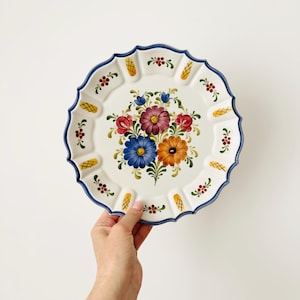 Op de afbeelding: Een ronde keramische bord met een geschulpte rand en een blauwe omlijning. Het midden toont een handgeschilderd bloemenboeket met paarse, blauwe en oranje bloemen. De rand is versierd met kleine rode bloemmotieven en gele ruitpatronen.