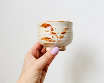 Puede incluir: Una pequeña taza de cerámica hecha a mano con una base de color crema y diseños marrones en forma de hojas. La taza tiene una forma redondeada y una pequeña base de pedestal. La taza se sostiene en una mano sobre un fondo blanco.