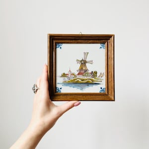May include: A framed ceramic tile with a Dutch windmill scene. The artwork shows a windmill, buildings, and water on a white background. The wooden frame has blue decorative corner accents.