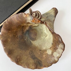 K&ouml;nnte beinhalten: Eine Keramikschale mit brauner und gr&uuml;ner Glasur. Die Schale hat eine einzigartige Form mit einer breiten &Ouml;ffnung und einem schmalen Boden. Die Schale ist mit einem kleinen, braunen Wirbel oben verziert.