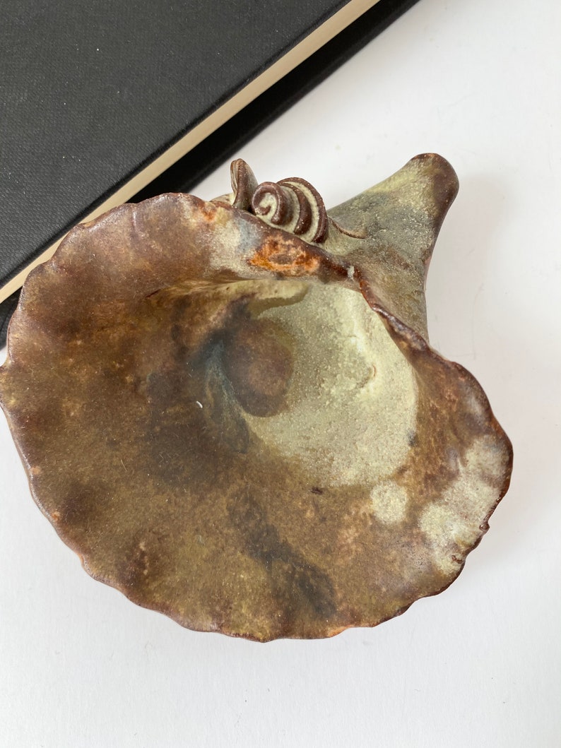 K&ouml;nnte beinhalten: Eine Keramikschale mit brauner und gr&uuml;ner Glasur. Die Schale hat eine einzigartige Form mit einer breiten &Ouml;ffnung und einem schmalen Boden. Die Schale ist mit einem kleinen, braunen Wirbel oben verziert.
