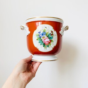 May include: A red ceramic planter with a white base and rim, decorated with gold trim. The planter has a central oval design with a colourful floral bouquet. Two ornate handles are on either side.
