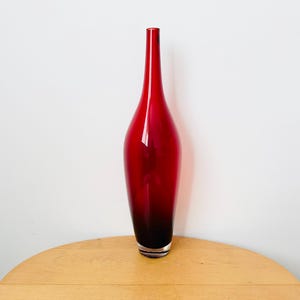 May include: A striking red glass vase with a long, slender neck and a rounded body. The vase features a colour gradient, going from a deep red at the top to a darker shade at the base. It is displayed on a wooden table against a white wall.