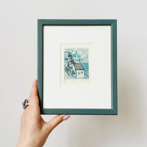 Könnte beinhalten: Ein gerahmter Aquarelldruck einer Küstenlandschaft. Das Kunstwerk zeigt weiße Gebäude mit roten und blauen Dächern, ein türkisfarbenes Meer und einen blau-grünen Rahmen. Der Druck wird von einer Hand gehalten.