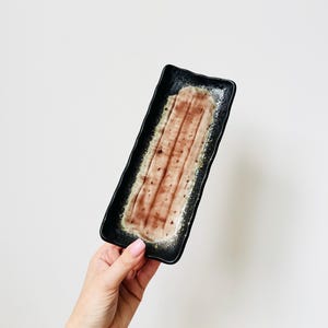 Könnte beinhalten: Ein rechteckiger Keramik-Servierteller mit schwarzer Außenseite und braun-beigem Innenraum. Der Teller hat eine strukturierte Oberfläche. Ideal zum Servieren von Vorspeisen oder kleinen Gerichten. Der Teller wird von einer Hand vor weißem Hintergrund gehalten.