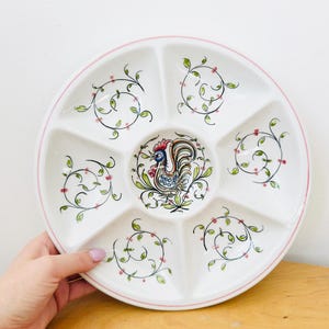 May include: White ceramic serving platter with six compartments and a pink rim. The center features a colorful rooster illustration. Each compartment is decorated with green leaves and pink berries.