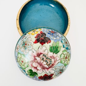 May include: A round, open trinket box with a floral design. The lid features a large pink peony surrounded by smaller flowers in red, yellow, and blue. The interior is a solid teal color, and the box has a gold-colored rim.