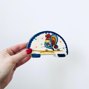 Puede incluir: Un reposacucharas de cerámica en forma de media luna con un diseño de gallo pintado a mano. El gallo presenta detalles azules, naranjas y amarillos sobre un fondo crema con detalles azules y naranjas. El reposacucharas tiene un borde festoneado azul.
