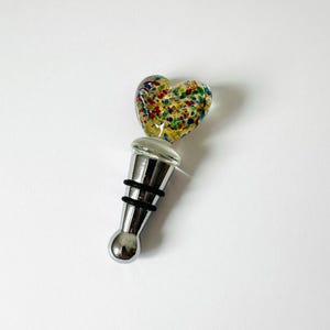 May include: A bottle stopper featuring a heart-shaped glass topper with a clear base and multicolored confetti-like flecks in red, blue, and green. The stopper has a polished silver-tone metal body with two black rubber rings for a secure seal.