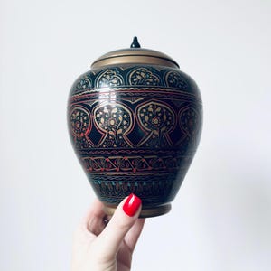 Puede incluir: Un jarrón decorativo con tapa, con base y tapa negras. El jarrón está adornado con intrincados patrones florales dorados, rojos y turquesas. La tapa tiene un pequeño pomo negro. El jarrón está sostenido por una mano con esmalte de uñas rojo.