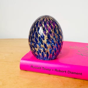 Puede incluir: Un pisapapeles decorativo de vidrio en forma de huevo. El pisapapeles presenta un diseño azul, dorado y morado. Está colocado sobre un libro rosa con el texto "Russell Tovey + Robert Diamant".