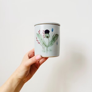 May include: A light gray ceramic jar with a lid, featuring a hand-painted floral design. The design includes a dark purple flower, green leaves, red berries, and blue accents. The jar is held by a person.