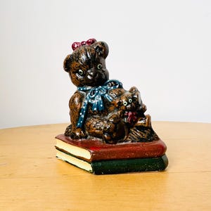Könnte beinhalten: Ein Keramik-Buchstütze mit einem braunen Teddybär mit einer blauen und weißen gepunkteten Schleife, der auf einem Stapel Bücher sitzt. Der Teddybär hält einen kleineren braunen Teddybär mit einer roten Schleife.