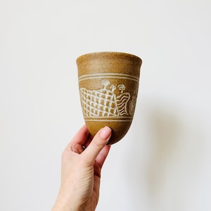 May include: A brown ceramic cup with a textured surface. The cup features a carved design of figures and a woven pattern. Two horizontal lines encircle the cup near the top. The cup is held by a hand.