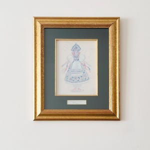 Könnte beinhalten: Ein gerahmtes Kunstwerk mit einer detaillierten Illustration eines traditionellen Kostüms. Die Zeichnung in Blau-, Rosa- und Weißtönen ist vor einem dunkelgrünen Passepartout und in einem goldfarbenen Rahmen gefasst. Das Kunstwerk ist an einer weißen Wand befestigt.