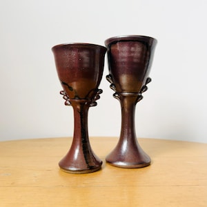 Può includere: Due calici in ceramica bordeaux scuro fatti a mano con finitura lucida. I calici hanno un bordo svasato, uno stelo decorativo e una base rotonda. Sono esposti su una superficie di legno chiaro.