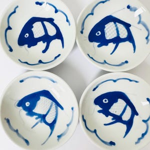 May include: Four small, white ceramic bowls with a blue fish design. Each bowl features a stylized blue fish in the center, surrounded by a blue wave-like border. The bowls are arranged in a square formation.